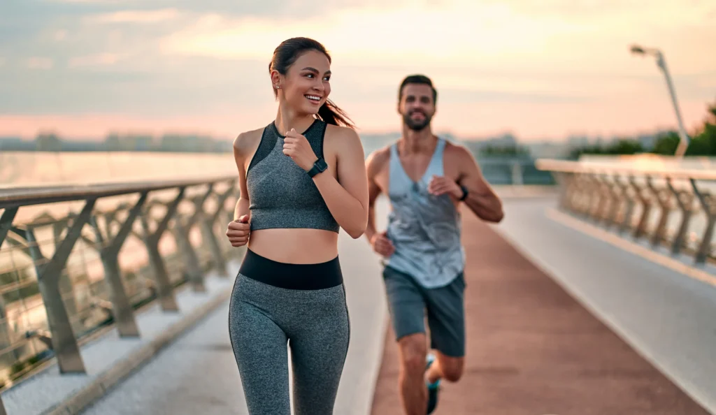 Two people jogging on a waterfront path during sunset wearing athletic clothing