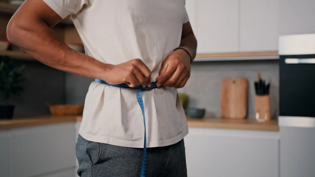 Person measuring their waist with a tape measure while standing in a kitchen